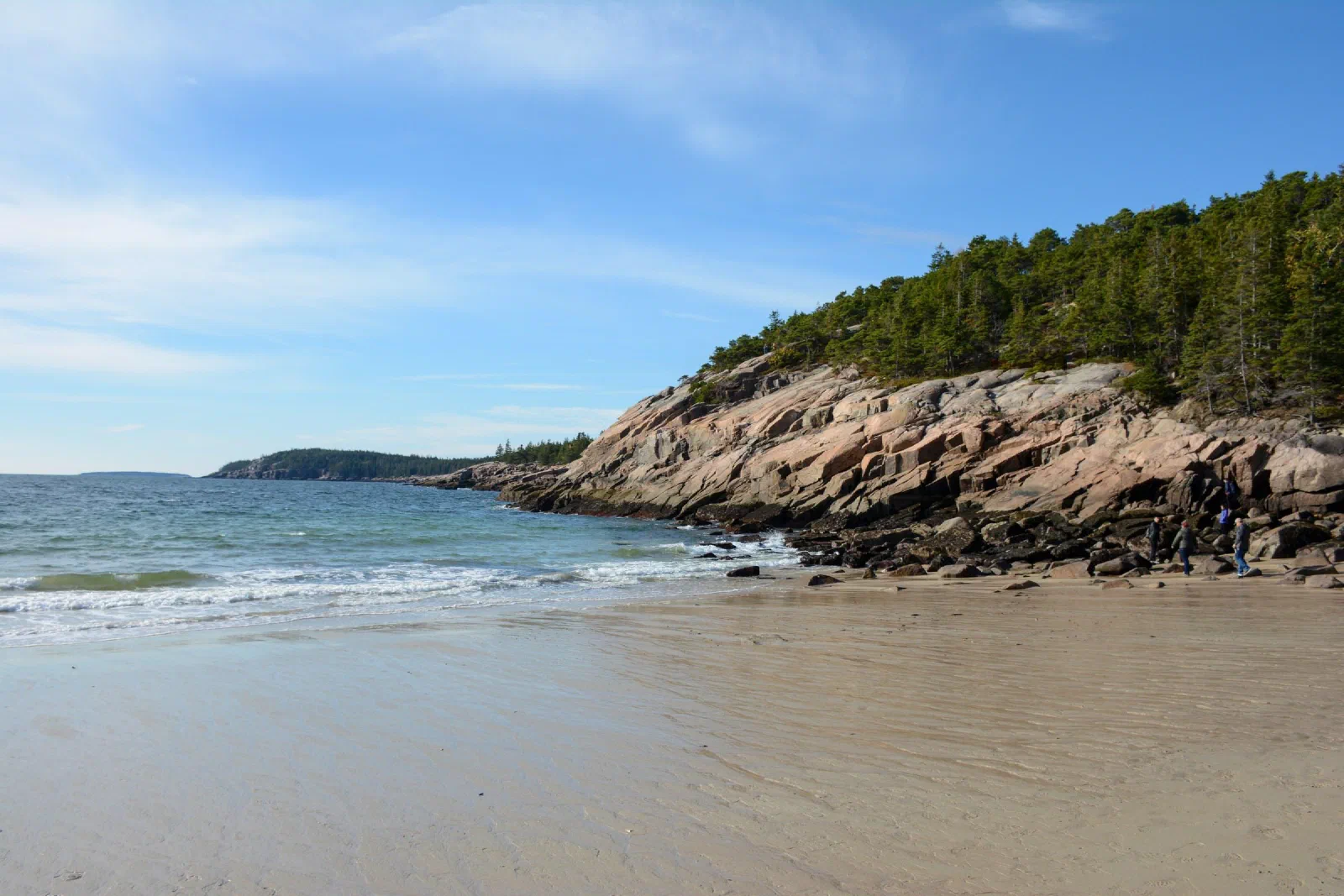 Small waves reaching a sandy beach beside a rocky, tree-lined shore.