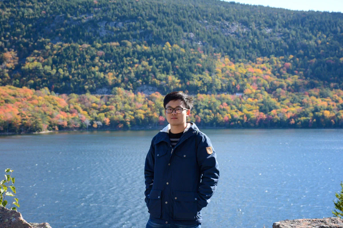 A person standing beside a lake with autumn-colored trees on the opposite shore.