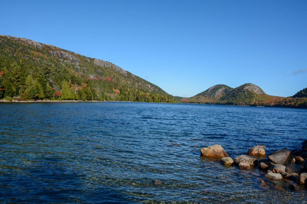 A blue lake bordered by rocks and wooded hills beneath a clear sky.