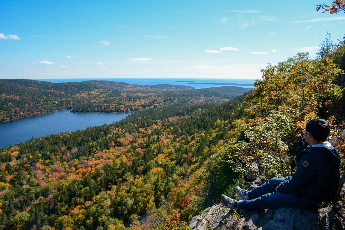 A person seated on a rocky overlook above a lake and hills covered in autumn foliage.