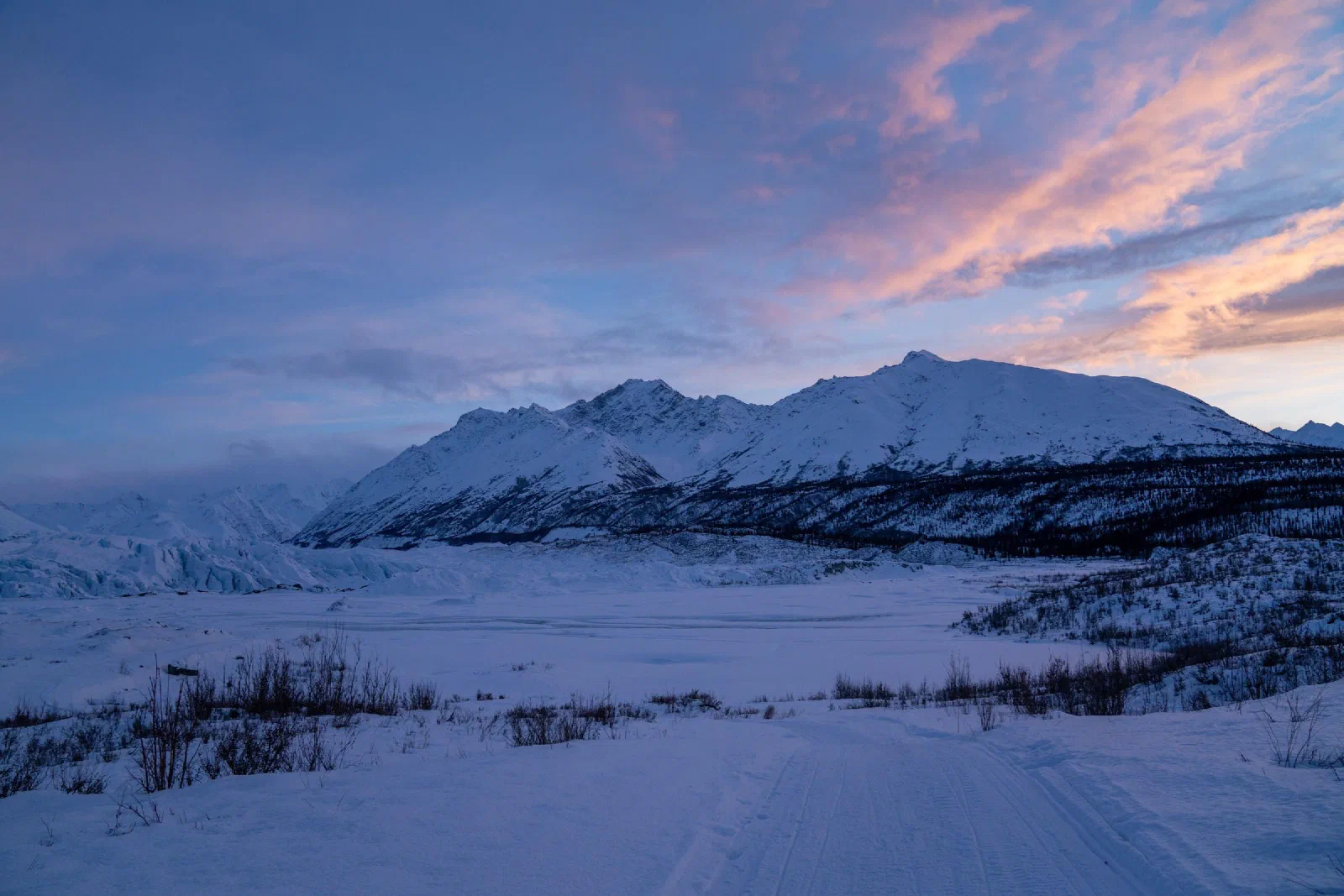 Snow-covered mountains beneath pink clouds, with a frozen valley in the foreground.