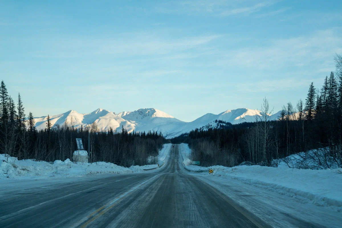 A straight snow-lined road leading toward distant mountains between evergreen trees.