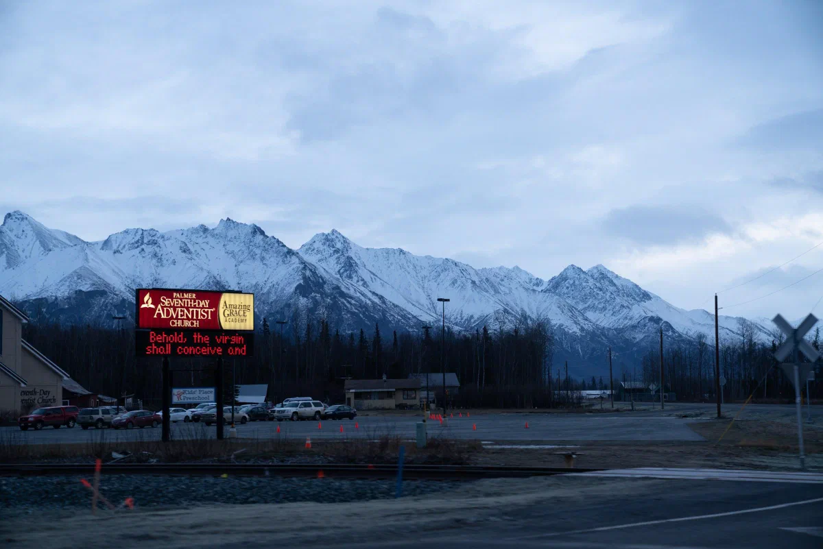 Snow-covered mountains behind a roadside sign and buildings beneath a cloudy sky.