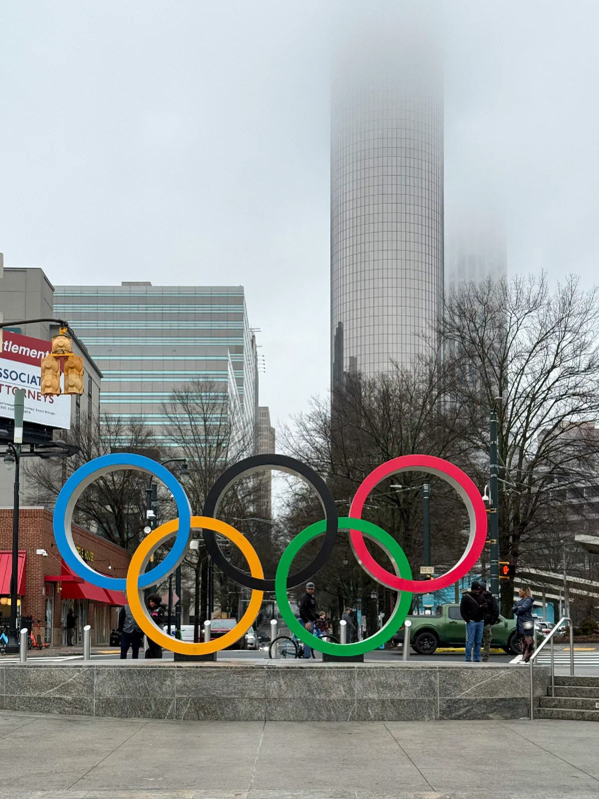 Five colored Olympic rings in front of trees and a tall building in the mist.