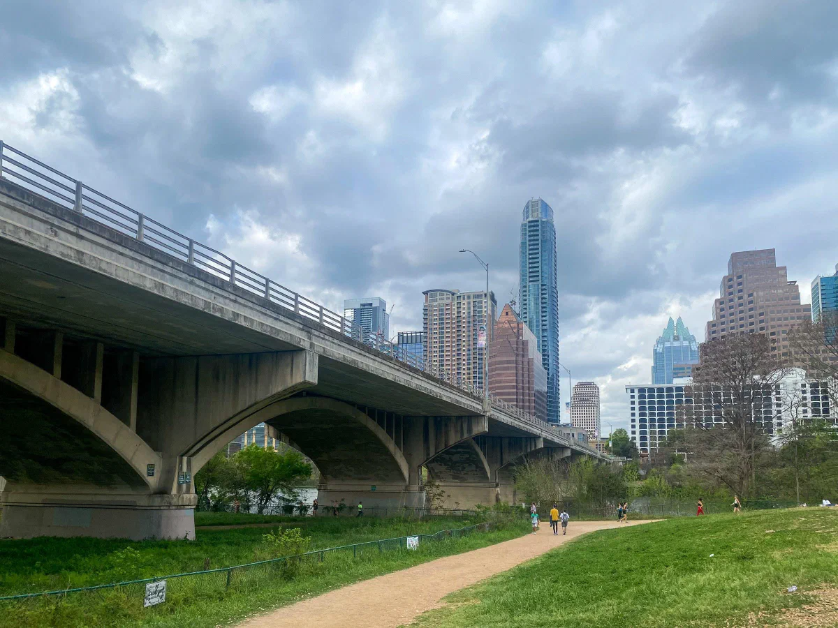 A concrete bridge beside a riverside path, with tall city buildings beyond.