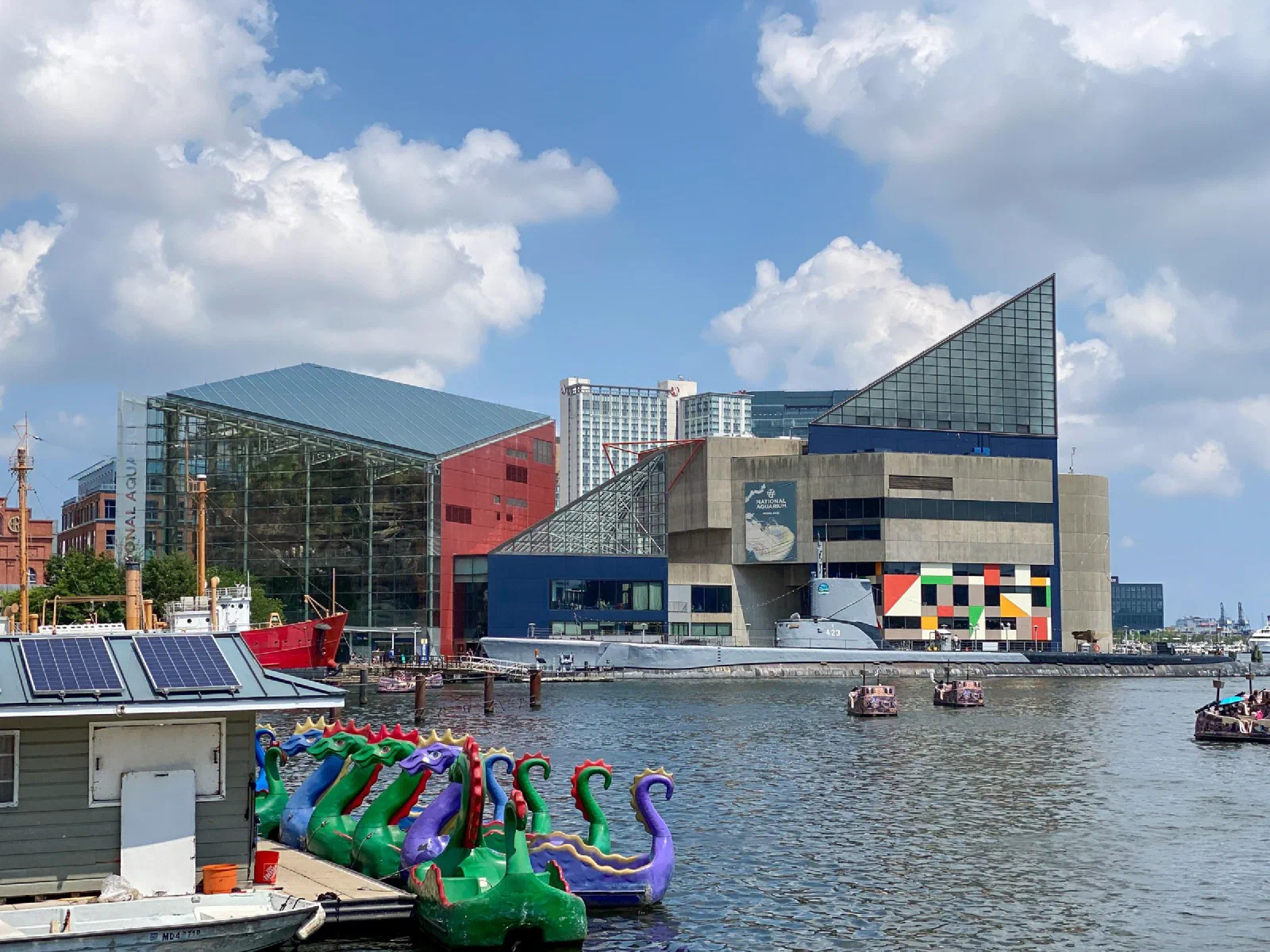 Boats and colorful pedal craft on the water in front of angular waterfront buildings.