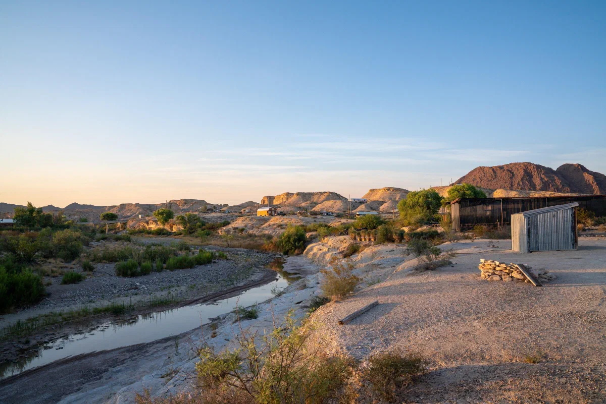 A narrow river winding through a dry landscape with low buildings and distant mountains.