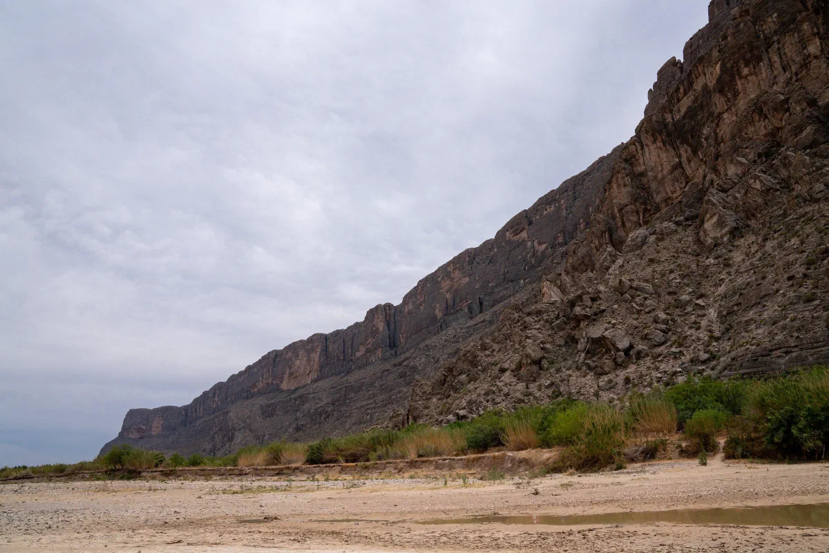 A sandy riverbank beneath a steep rocky canyon wall and an overcast sky.