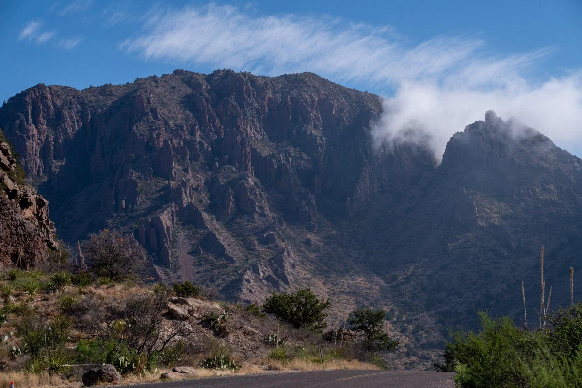 Clouds brushing the top of a steep rocky mountain above desert shrubs.