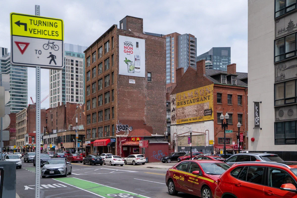 Cars passing brick buildings and restaurant signs along a city street.