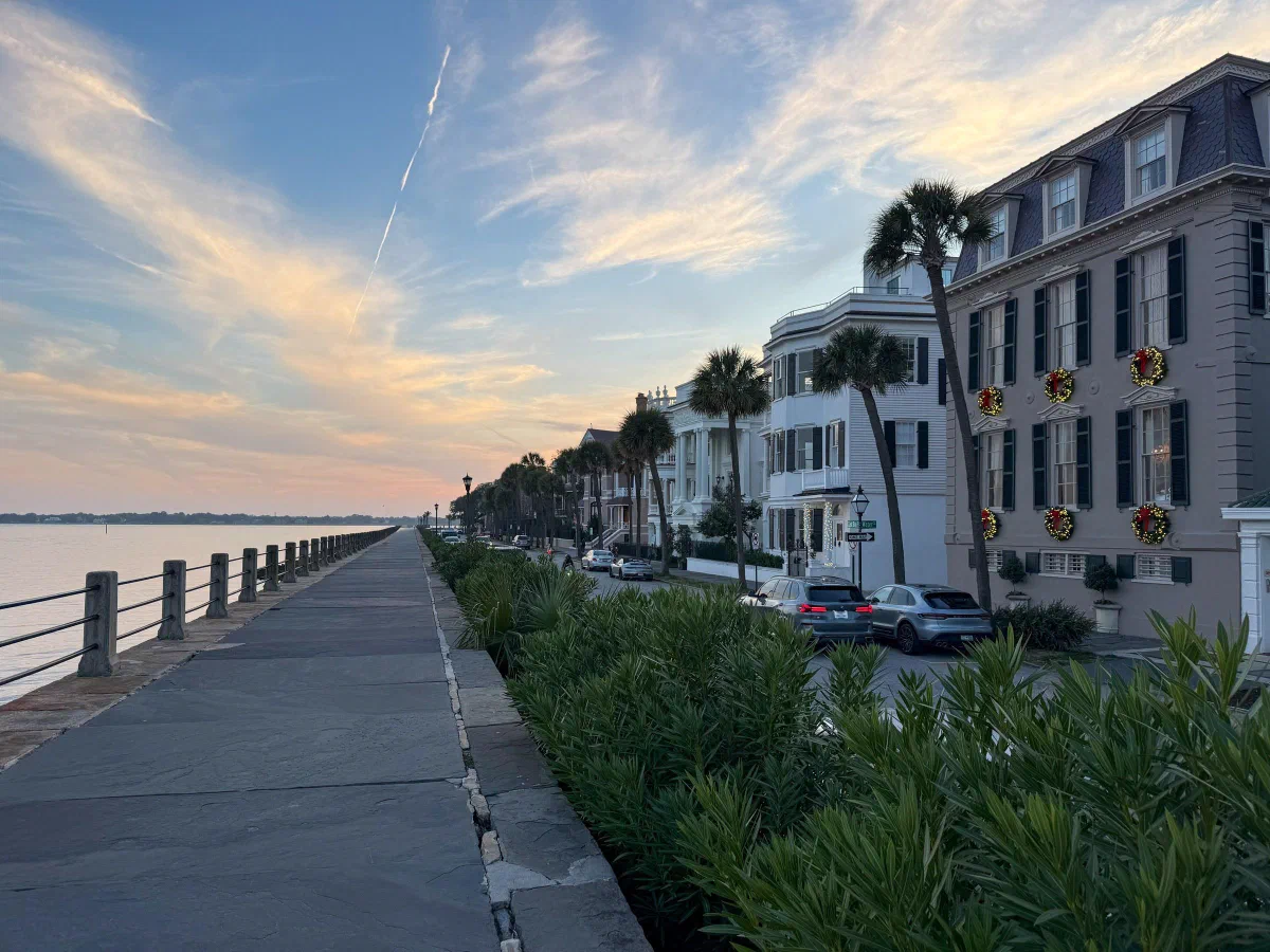A waterfront path beside tall houses and palm trees under a colorful evening sky.
