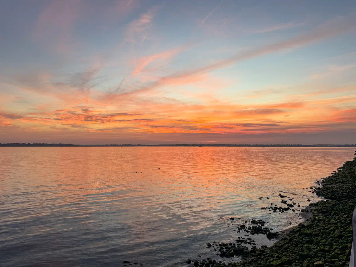 Orange and pink sunset clouds reflected in calm water beside a rocky shore.