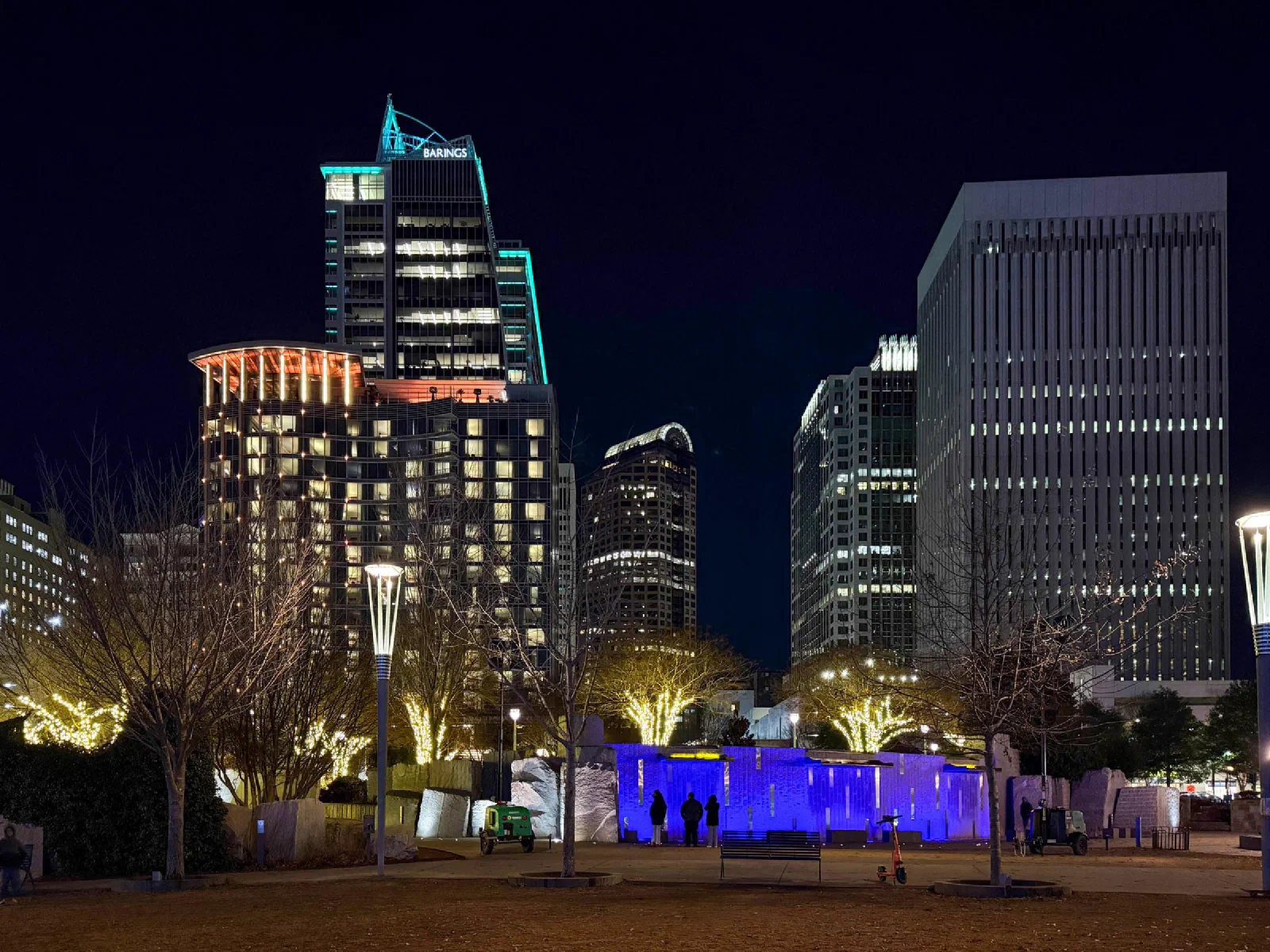Illuminated towers rise above trees wrapped in lights and a blue-lit stone wall at night.