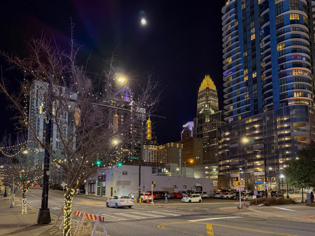 A moonlit street intersection with trees wrapped in lights, a rounded high-rise, and a tower with a golden crown.