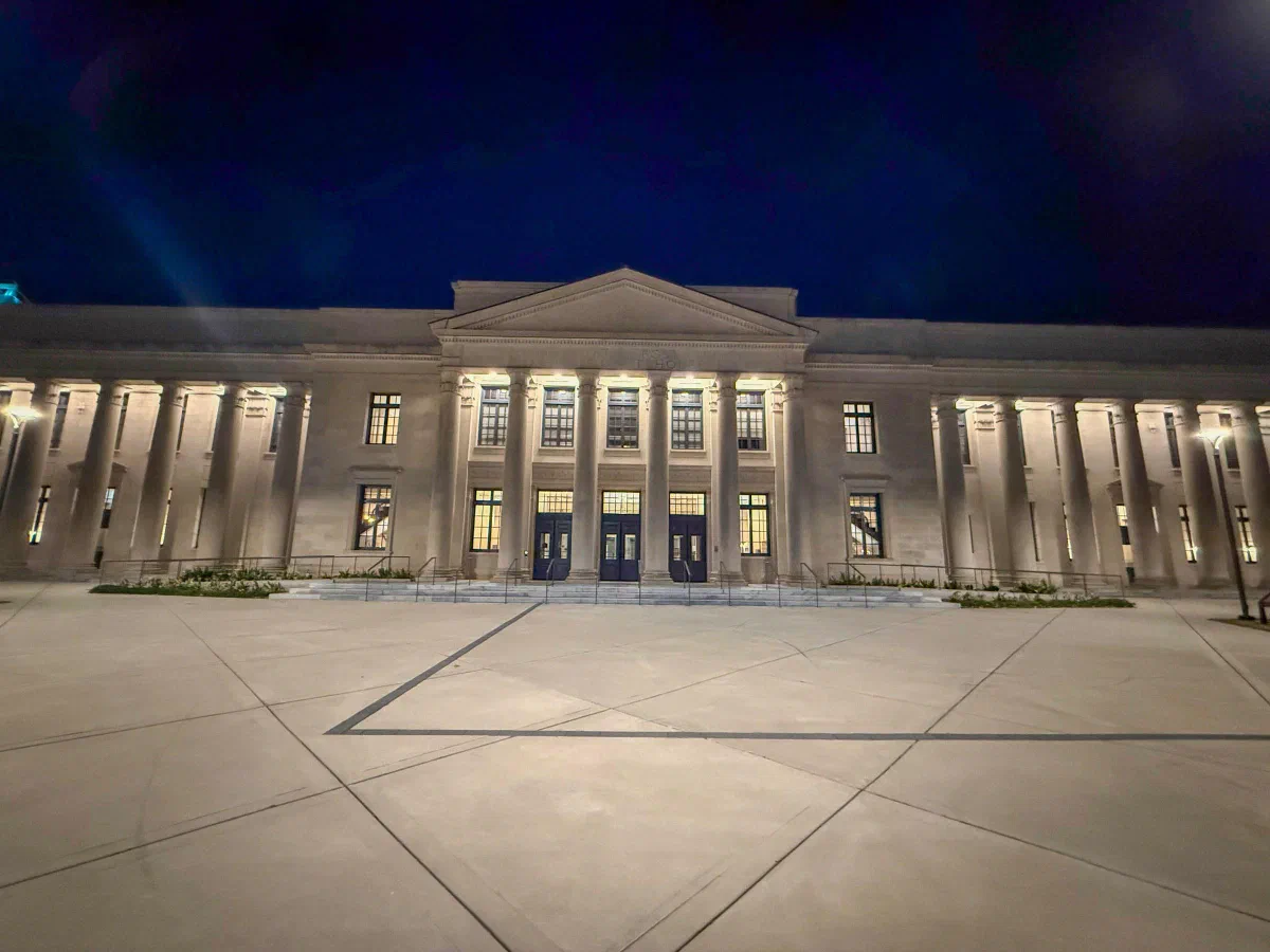 An illuminated stone building with tall columns and a broad paved plaza beneath a dark blue evening sky.