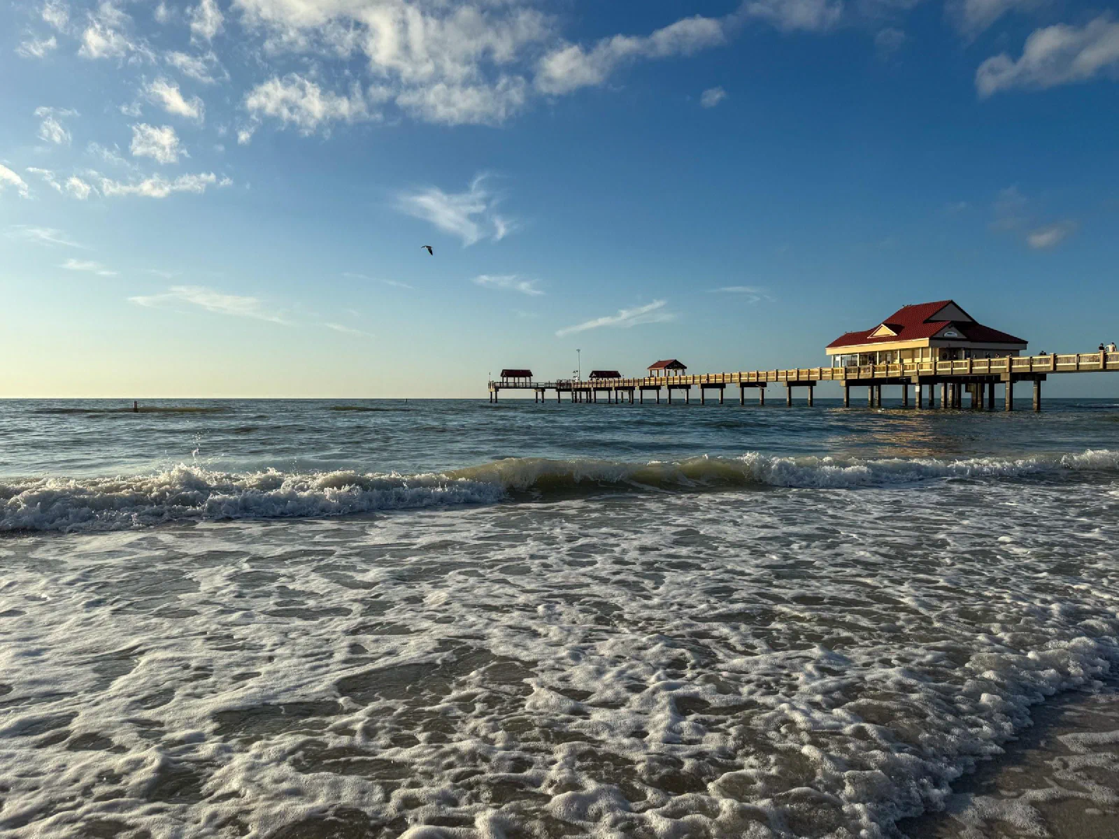 Waves rolling toward a sandy beach beside a long pier under scattered clouds.