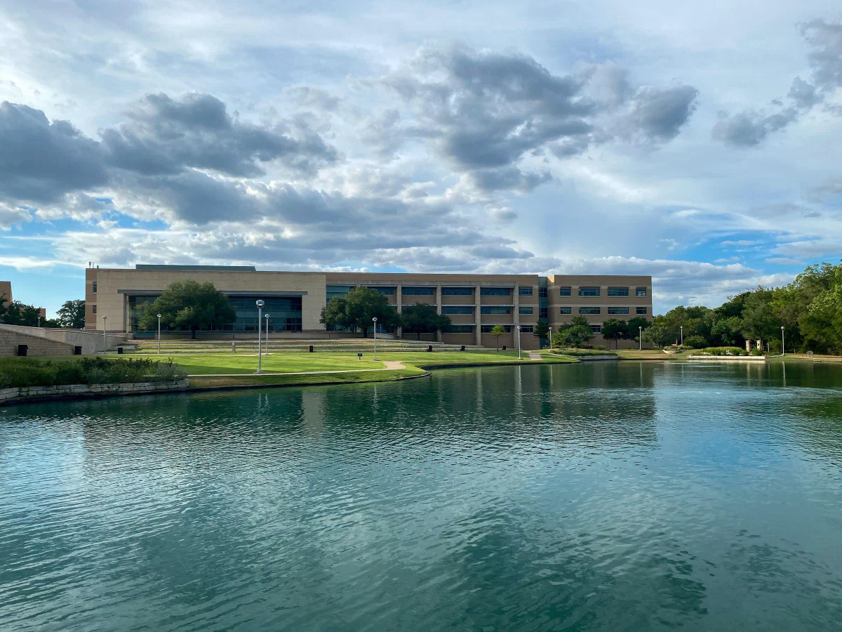 A broad campus building across a lake, with trees and dramatic clouds overhead.