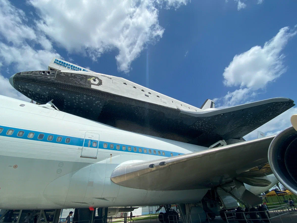 A space shuttle mounted above a large aircraft beneath a bright blue sky.