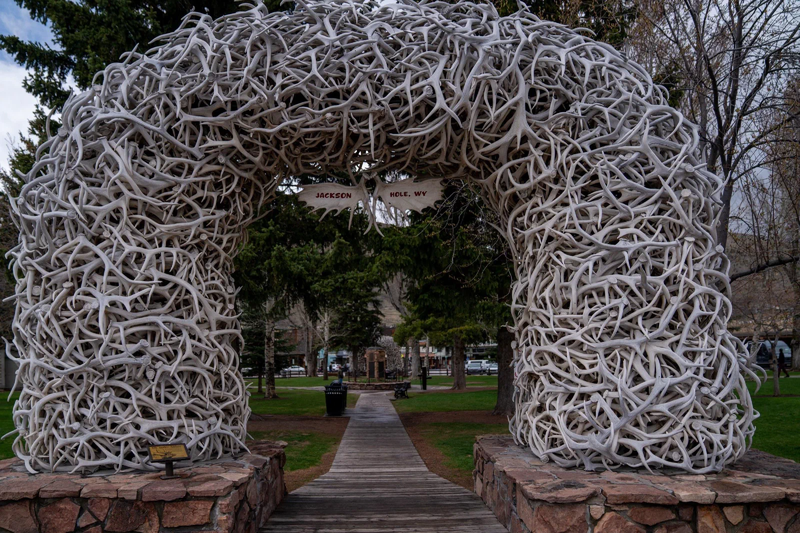 An arch made of interwoven antlers above a paved entrance to a tree-filled park.