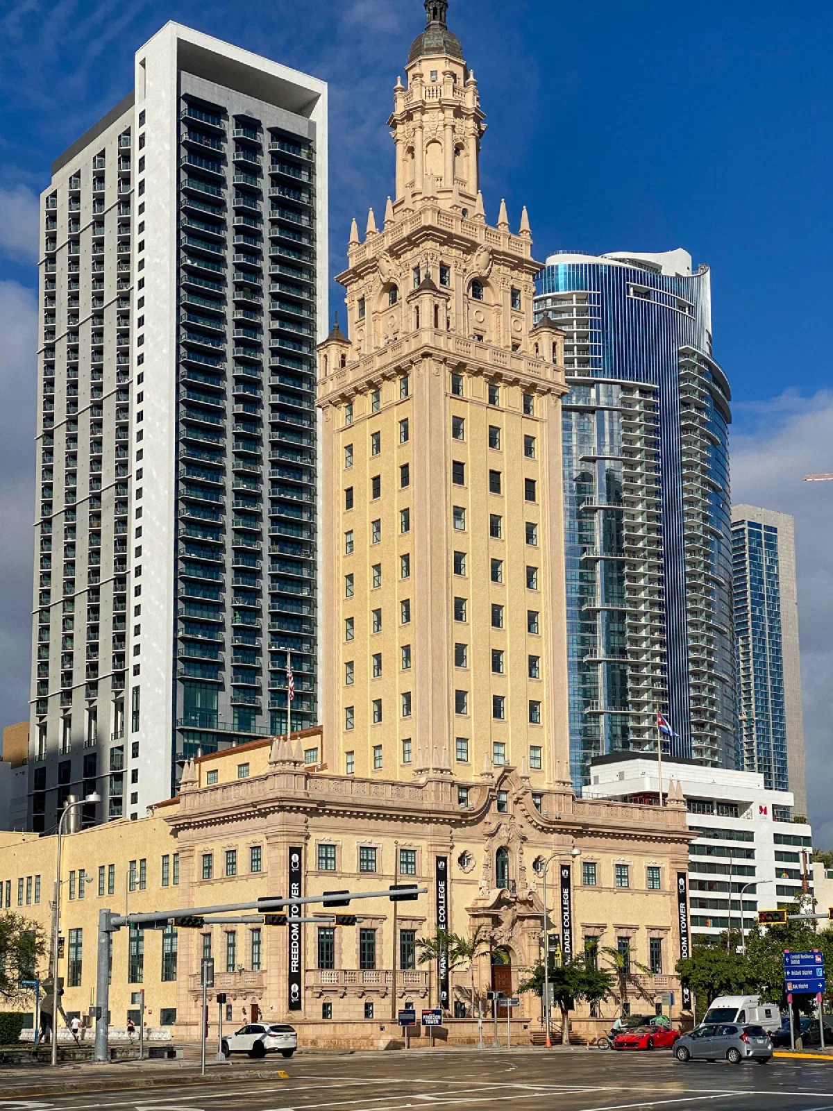 A pale ornate tower in front of taller modern buildings beside a busy street.