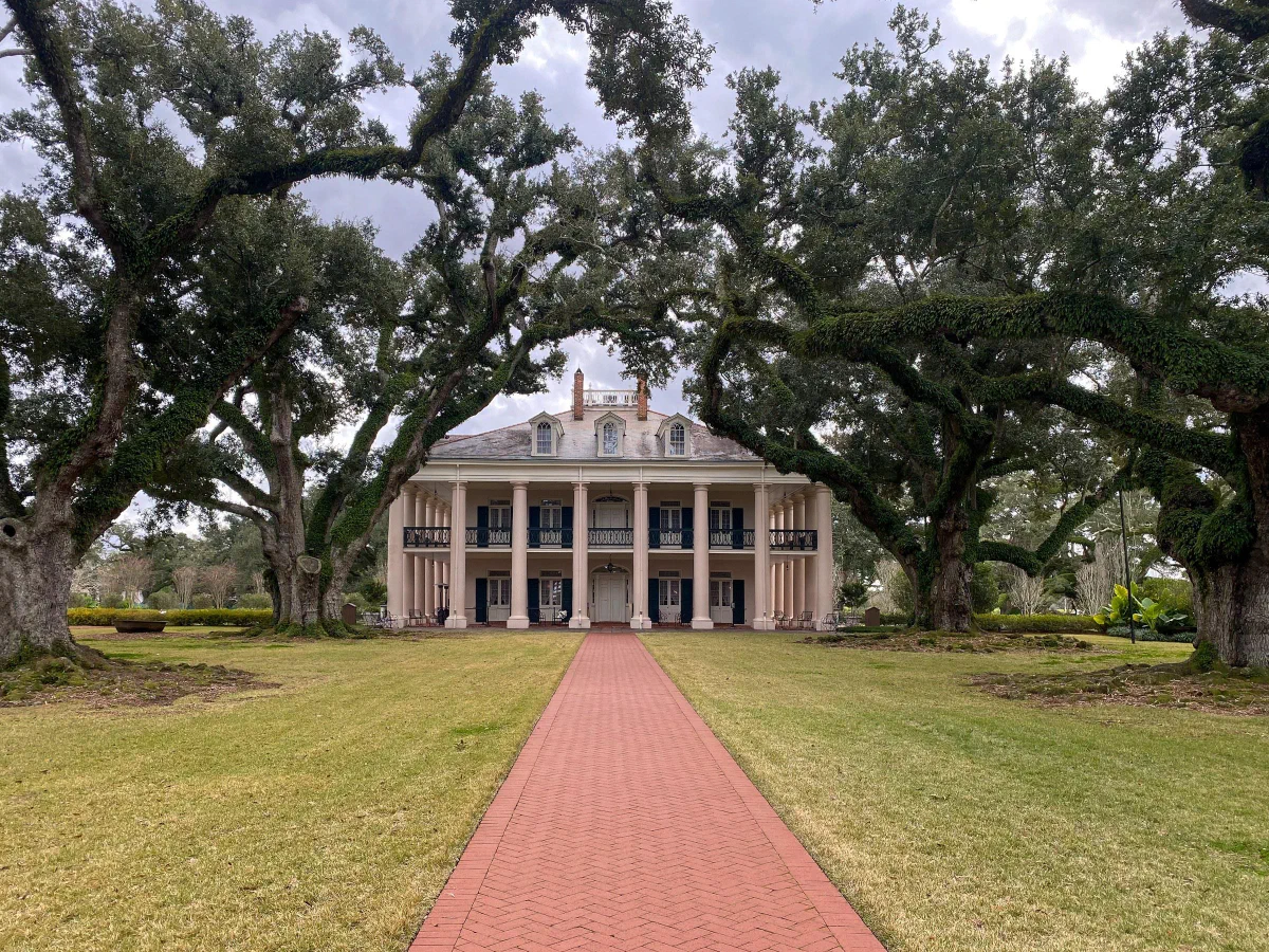A columned house at the end of a red brick path framed by spreading oak trees.