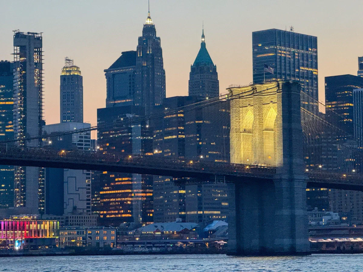 An illuminated suspension bridge in front of the city skyline at dusk.