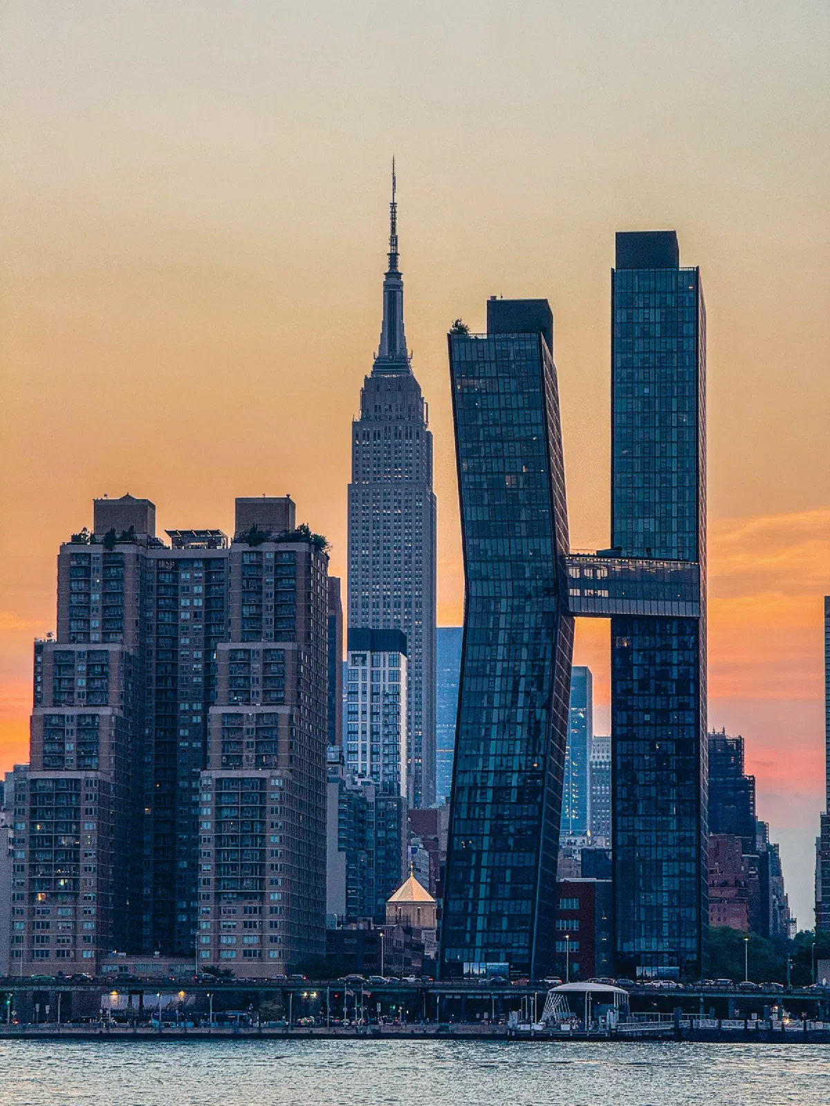 Tall waterfront buildings silhouetted against a warm orange evening sky.