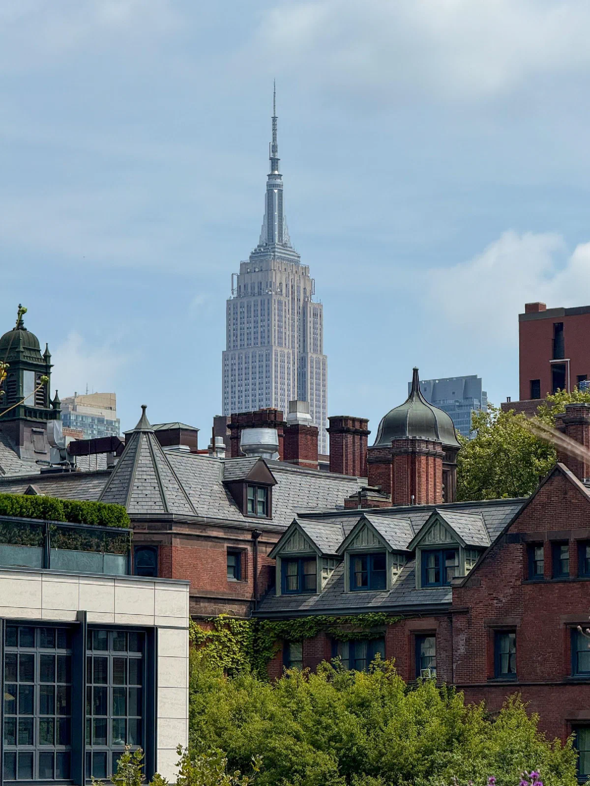 A tall spired skyscraper rising beyond brick rooftops and leafy trees.