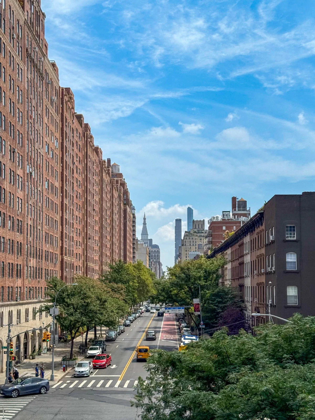 A long city street lined with brick buildings, viewed from above through green trees.