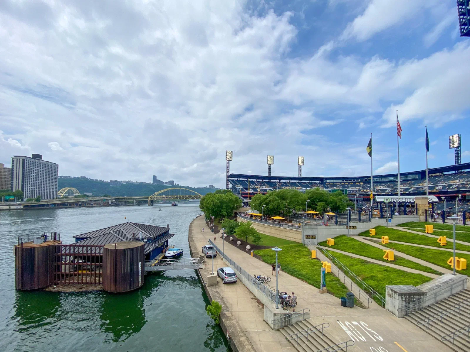 A riverside walkway beside a baseball stadium, with boats and yellow bridges beyond.