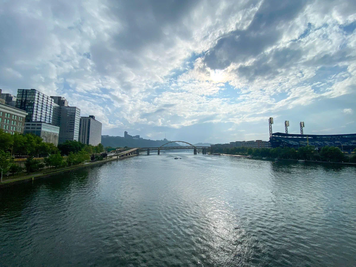 A broad river between city buildings and a stadium, with bridges beneath scattered clouds.