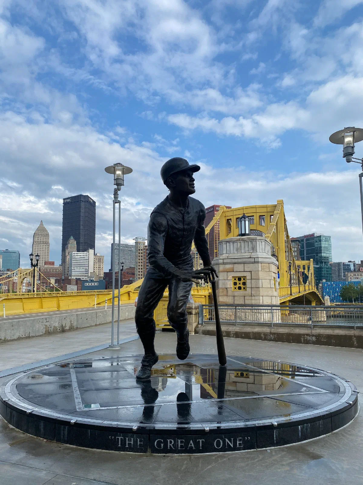 A bronze baseball player statue beside a yellow bridge, with city buildings beyond.