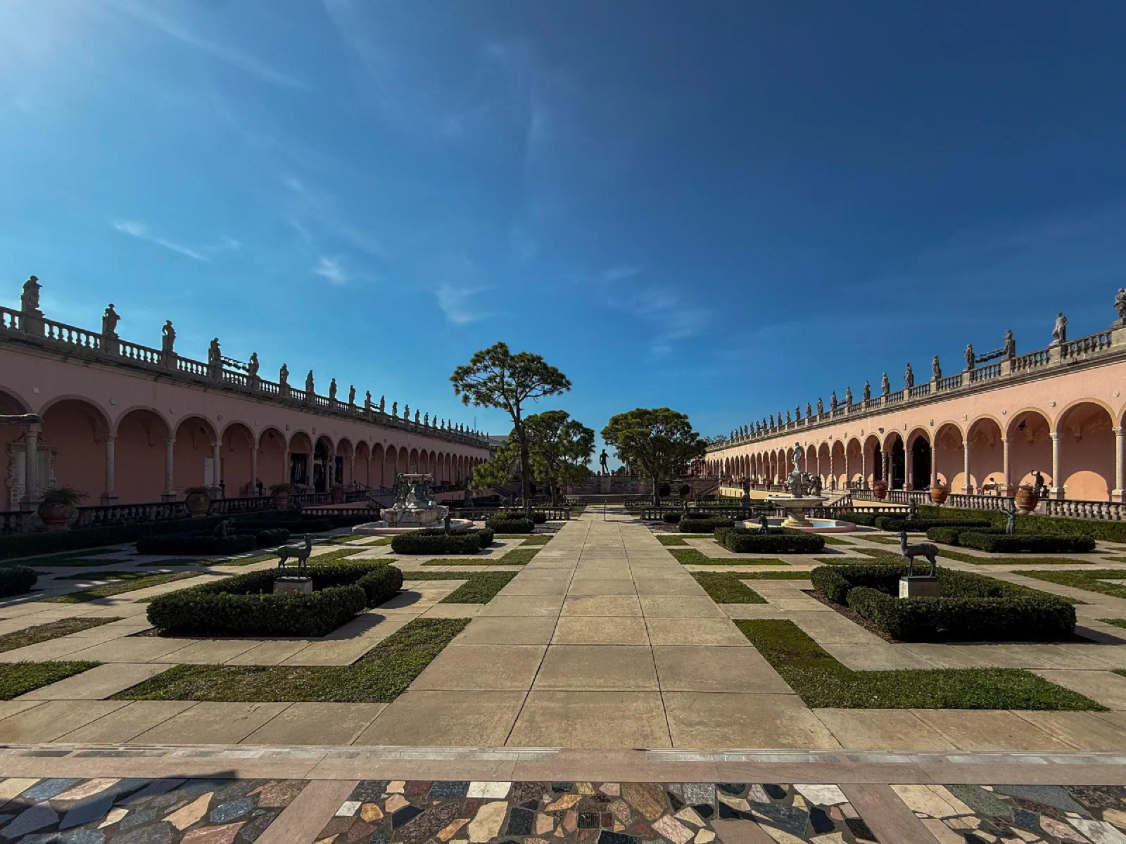 A formal courtyard with clipped hedges, a central walkway, and long pink arcades.