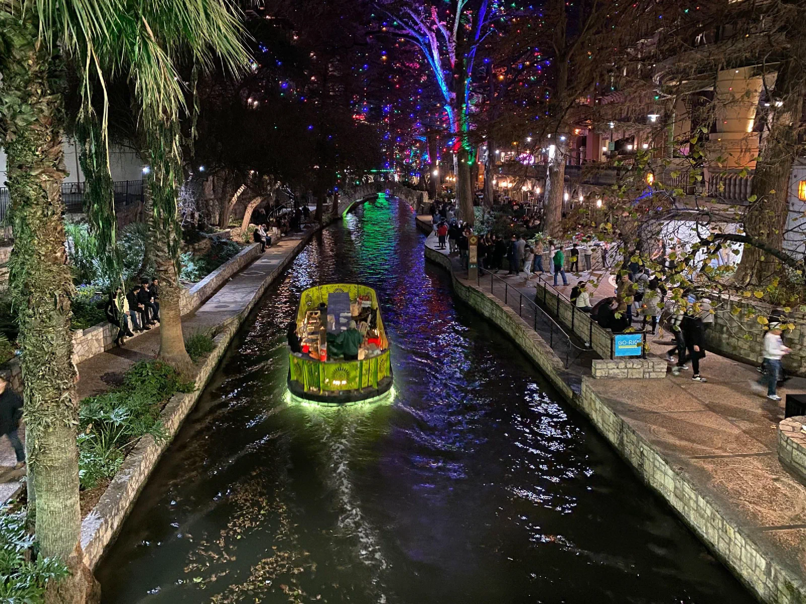 A brightly colored boat passing through a narrow canal lined with lights at night.