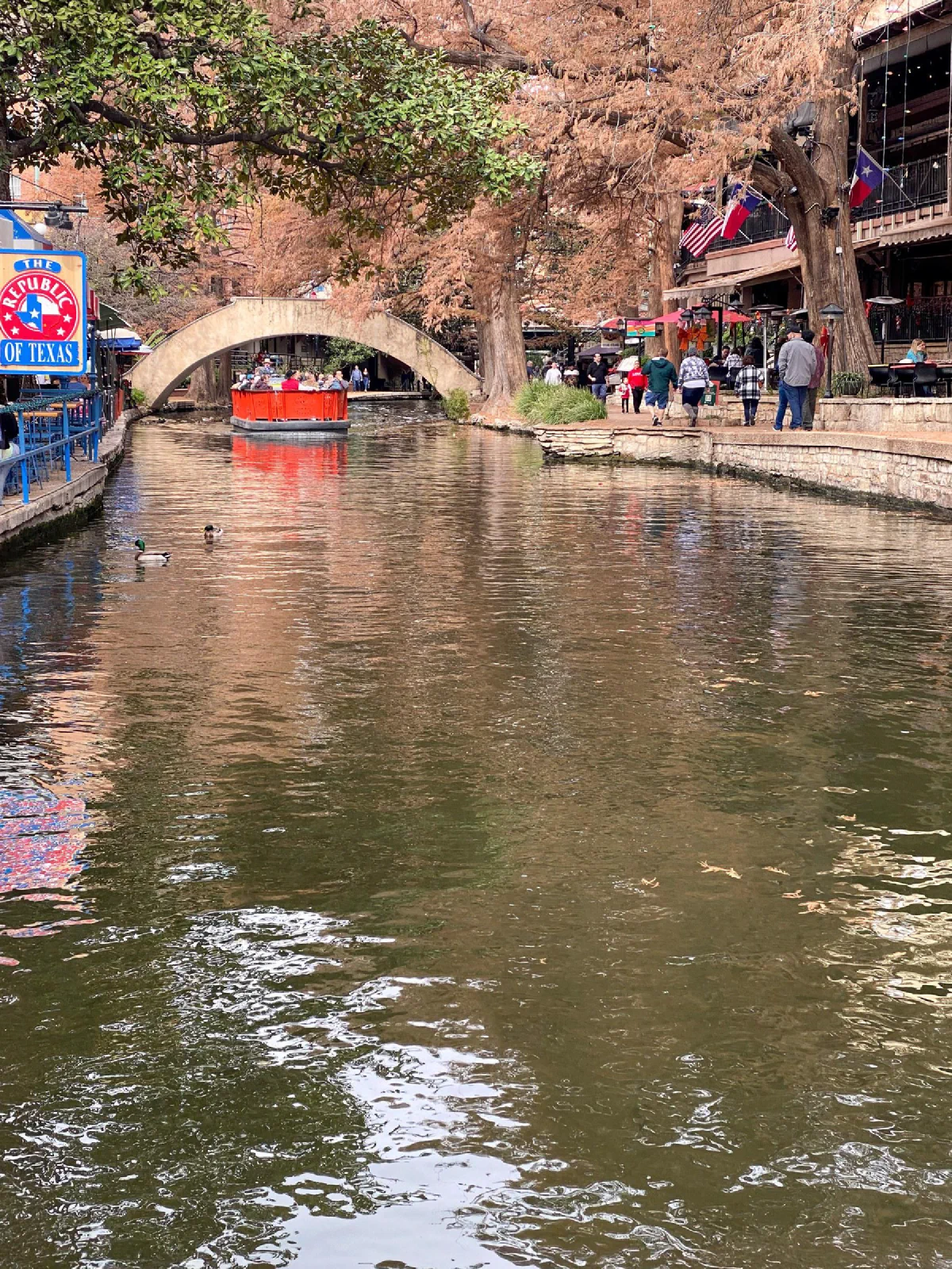 A red boat beneath a stone bridge on a canal bordered by walkways and buildings.