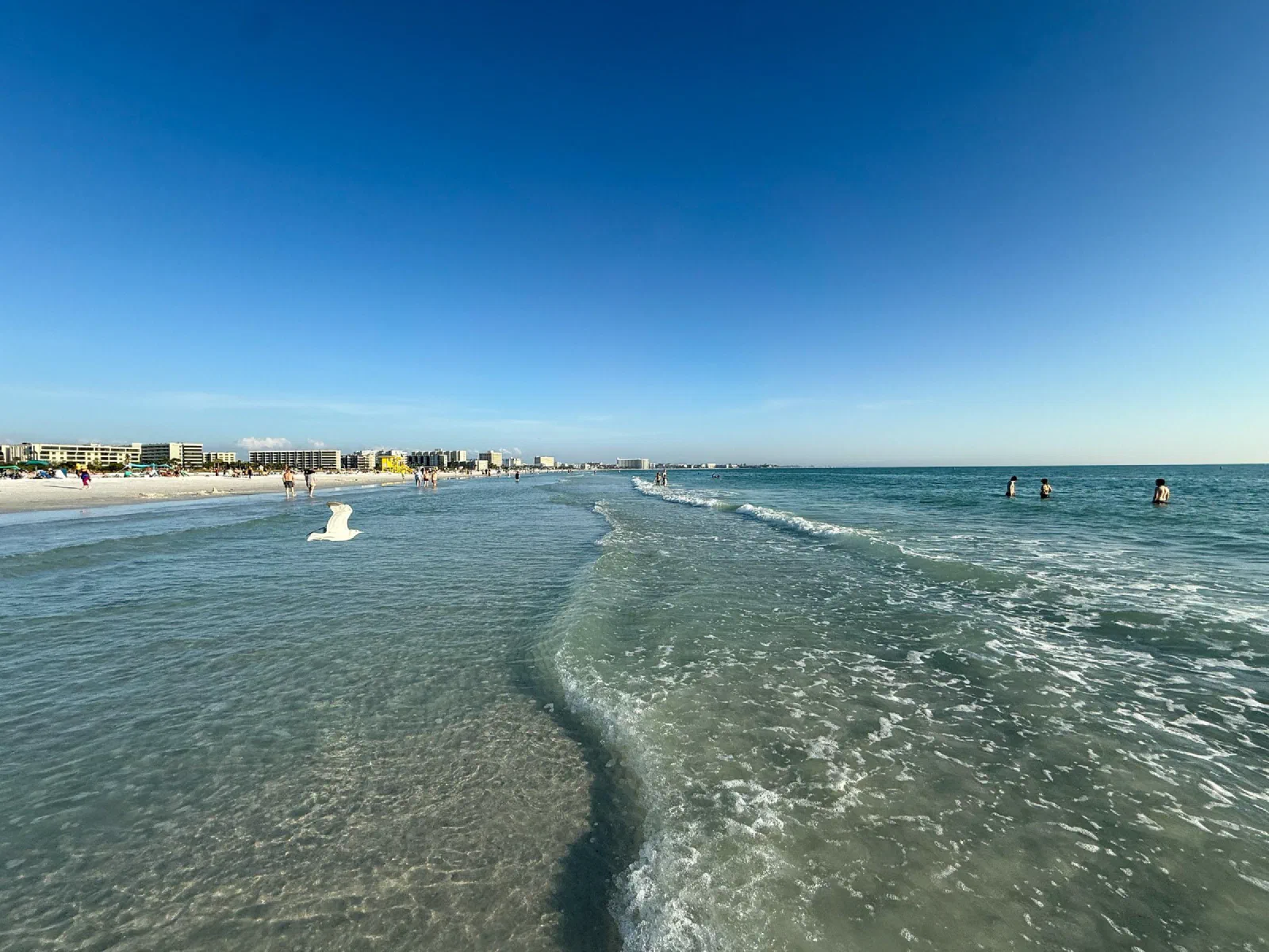 Small waves rolling onto a sandy beach with distant people and buildings along the coast.