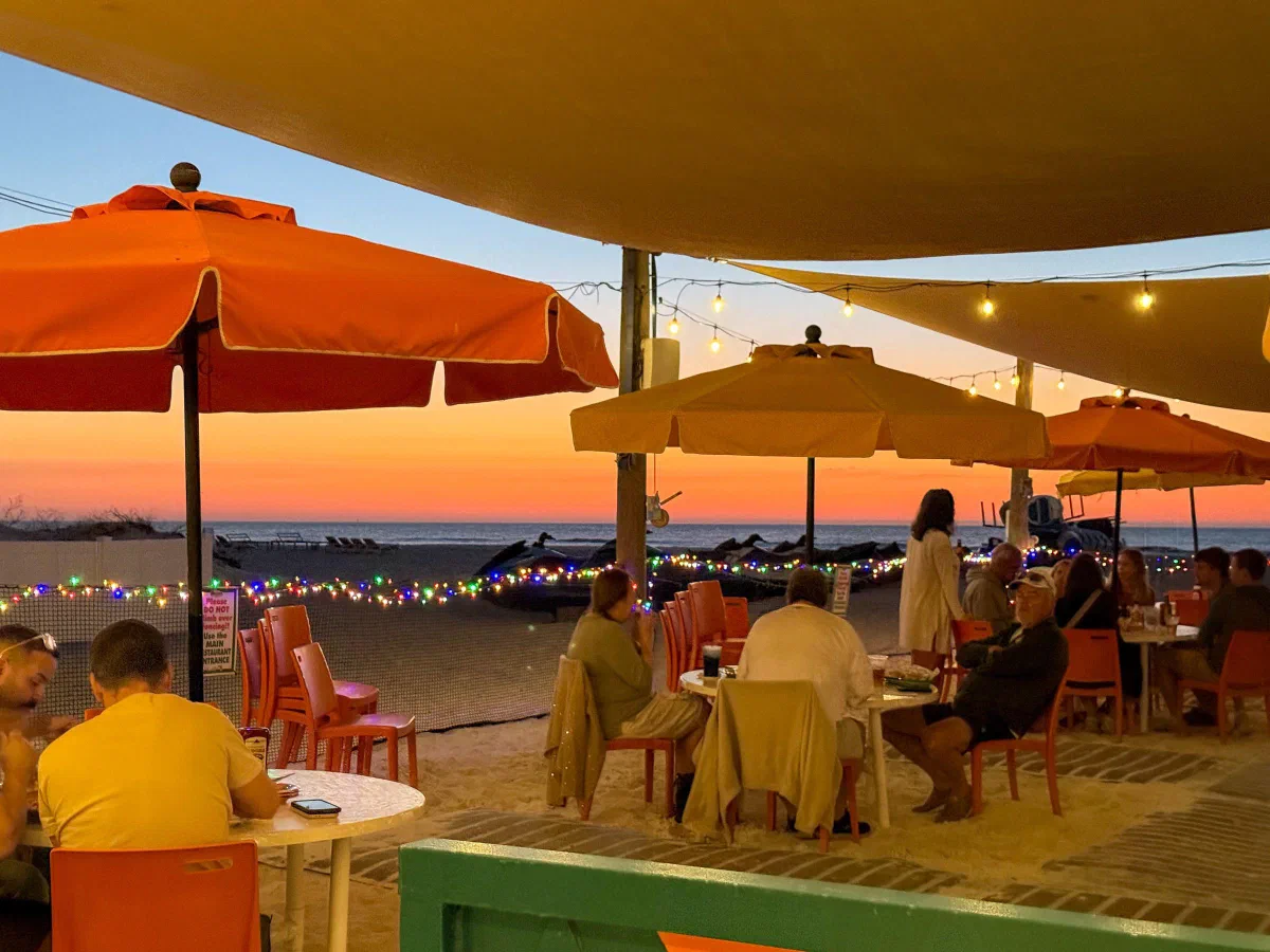People seated beneath brightly colored umbrellas at an outdoor terrace in evening light.
