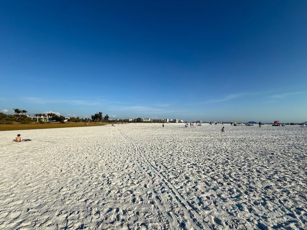A wide, pale sandy beach with footprints and distant beachgoers beneath a blue sky.
