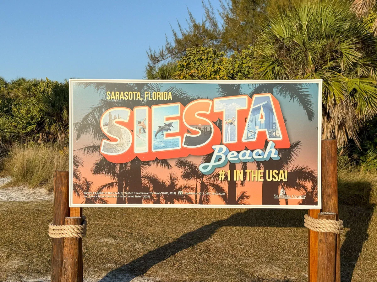 A colorful Siesta Beach sign beside sand, beach grass, and shrubs.