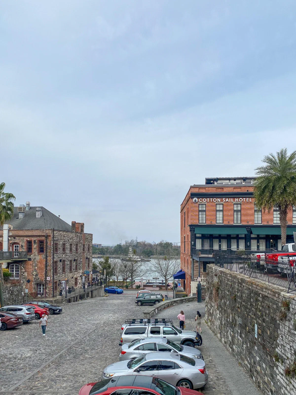 Parked cars beside a sloping brick street and brick buildings near the water.