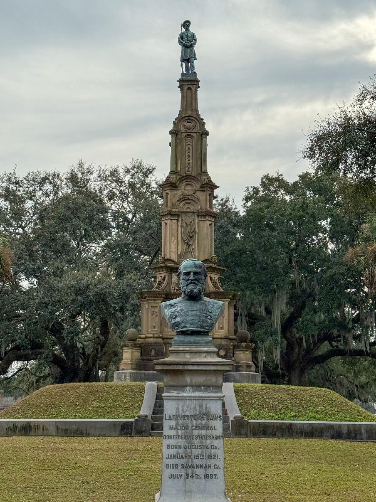 A tall carved stone monument and statues in a landscaped park beneath a cloudy sky.
