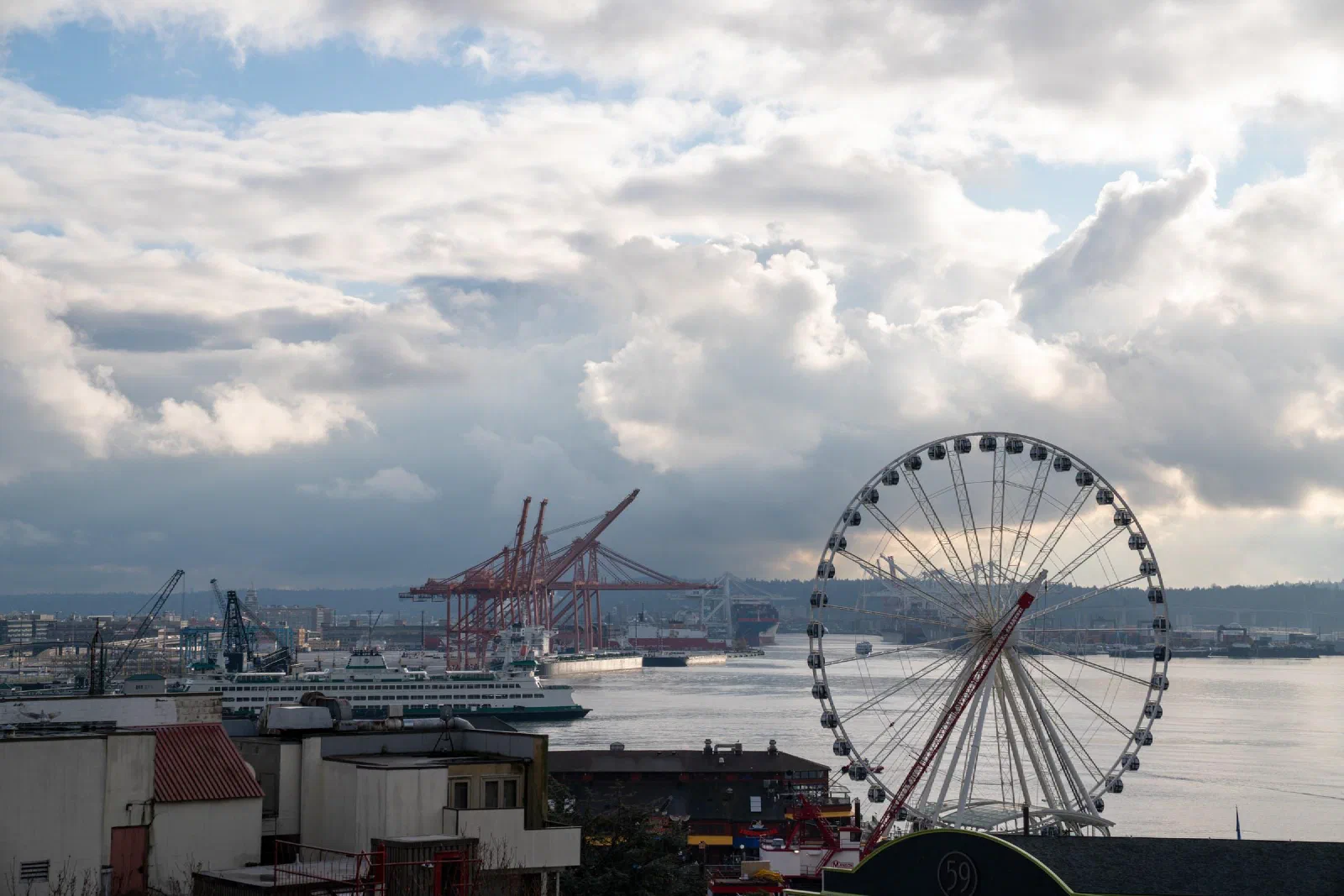 A Ferris wheel above the waterfront, with cranes beneath a cloudy sky.