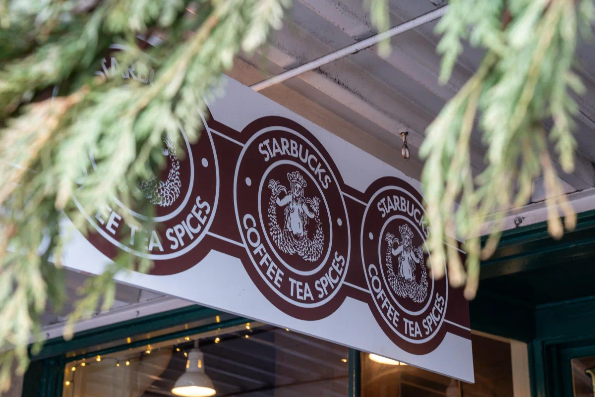 Brown Starbucks coffee signs hanging beneath an awning framed by evergreen branches.