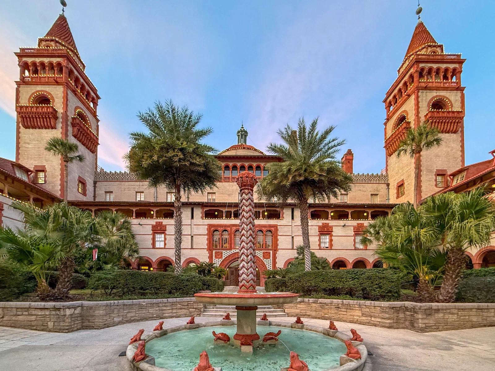 A fountain and red-roofed towers framed by palm trees and formal gardens.