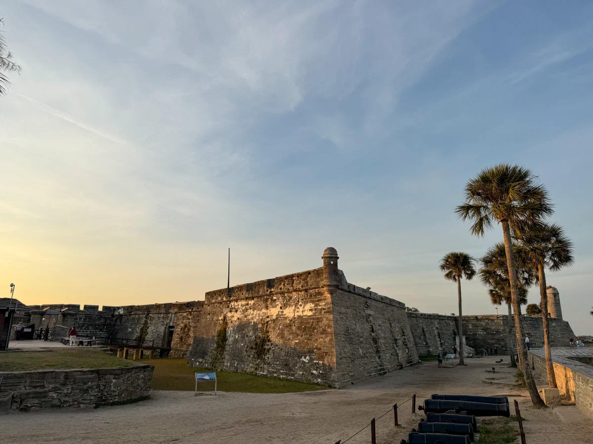 A low stone fort wall with a projecting corner tower and palm trees in warm light.