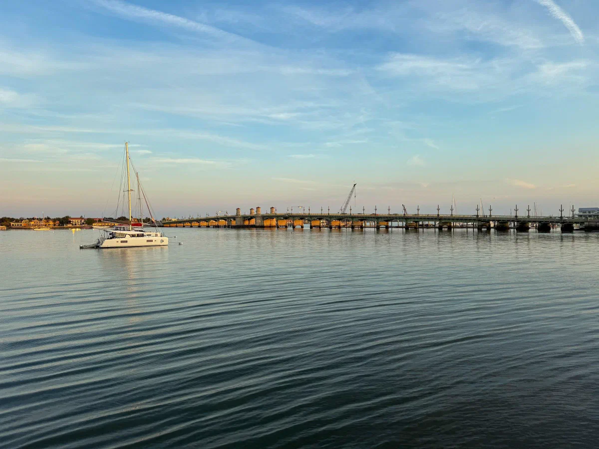 A sailboat on calm water near a long low bridge in the evening light.