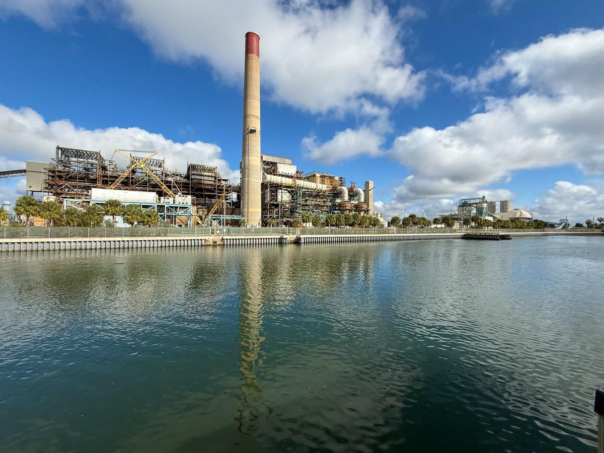 An industrial waterfront with a tall smokestack reflected in calm water.