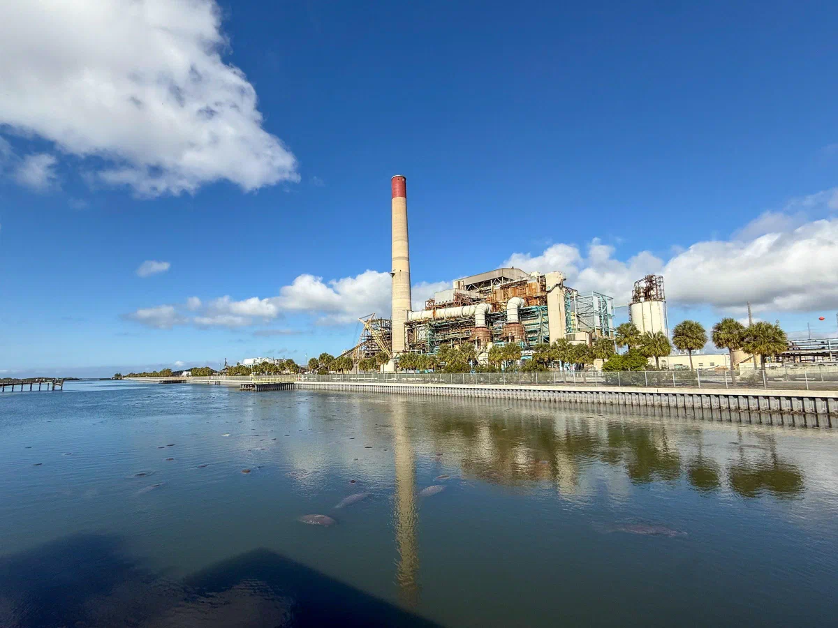 A waterside industrial complex and smokestack beneath white clouds.