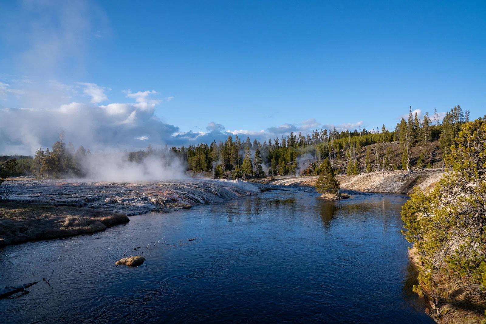 Steam rising beside a blue river bordered by pale rock and evergreen trees.